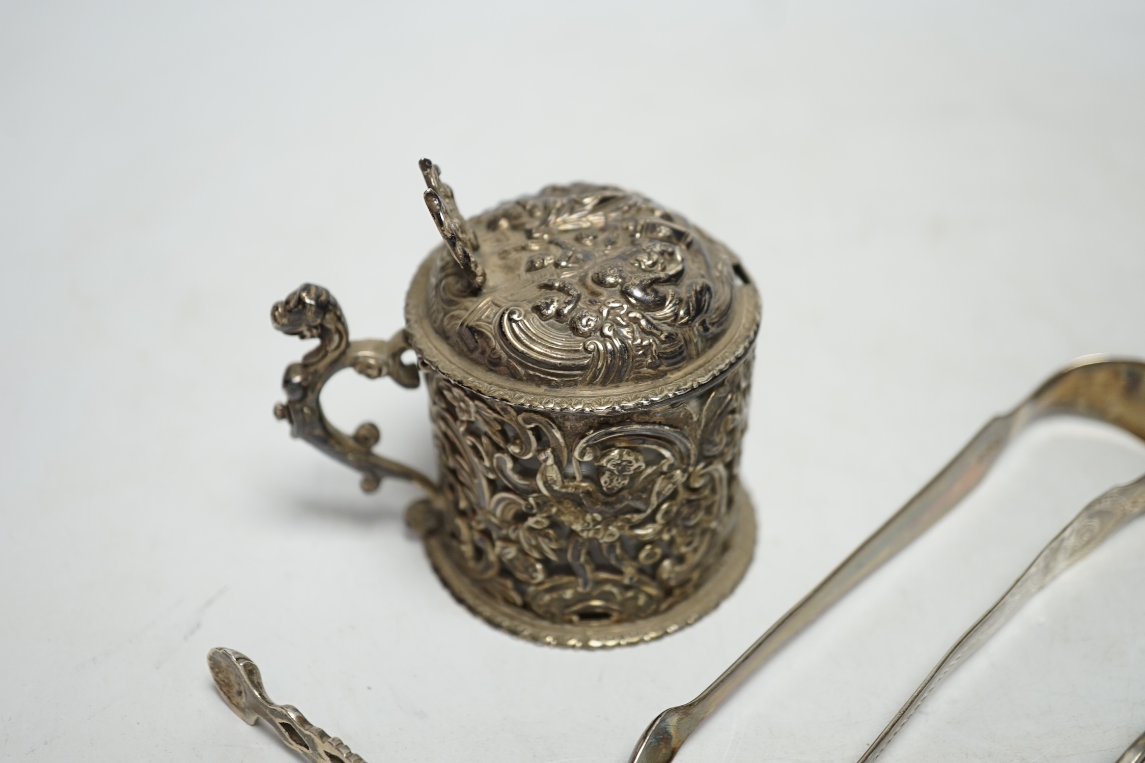 A Victorian silver embossed mustard pot, with spoon and three pairs of George III and later silver sugar tongs.